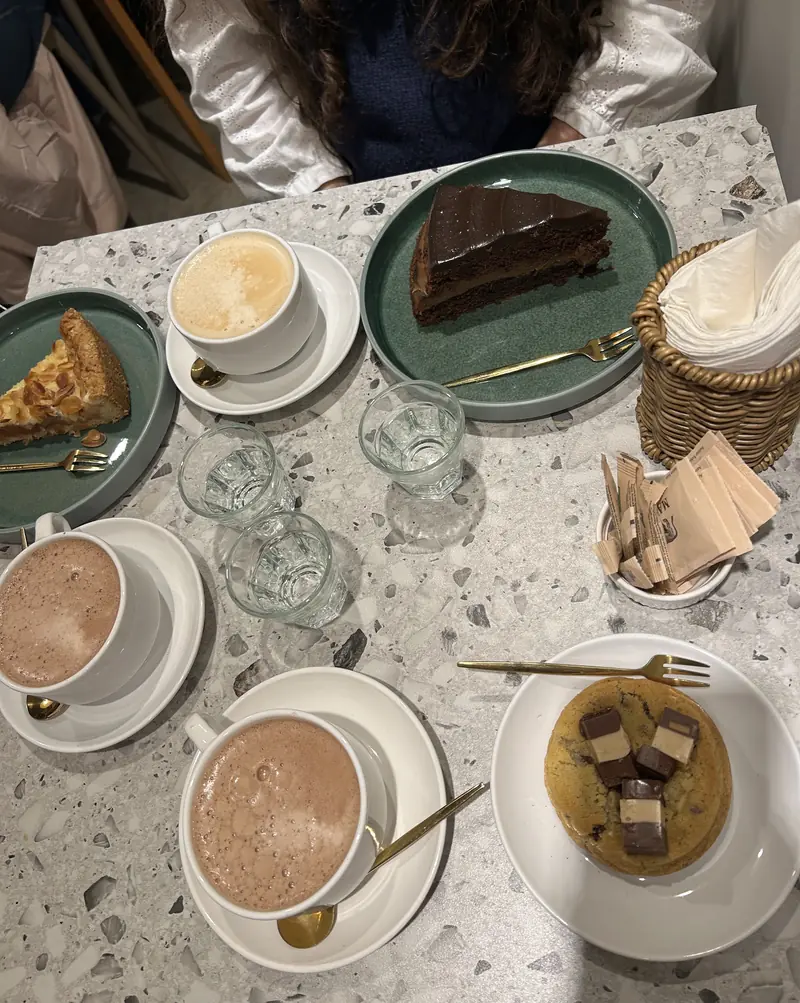 Vista desde arriba de una mesa de terrazzo con chocolate caliente, café, porciones de torta y una cookie