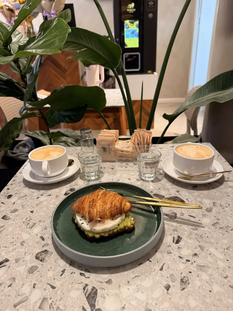 Mesa de terrazzo con dos cafés con leche y un croissant con palta, entre plantas, adentro de Lo de Anita